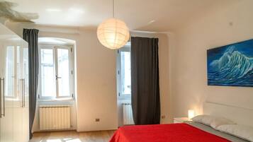 Een slaapkamer met een rode sprei, een schilderij van een golf, een hanglamp en uitzicht naar buiten door een raam.