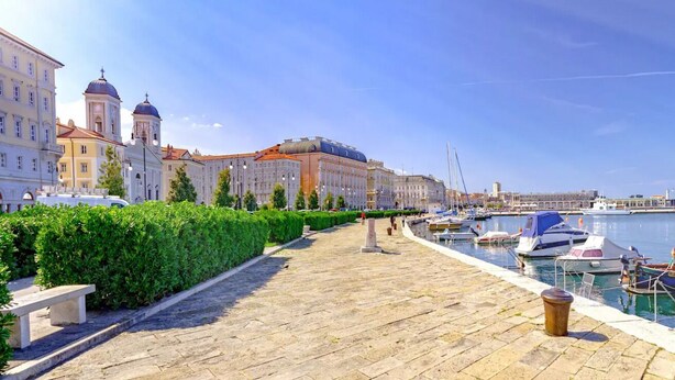 A waterfront promenade with a stone pathway, a row of buildings with domed architecture, and a marina with boats.