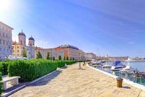 A waterfront promenade with a stone pathway, a row of buildings with domed architecture, and a marina with boats. - Hadithi Trieste, between Le Rive and Piazza Unità (Trieste)