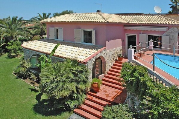 Eine pinkfarbene Villa mit einem Ziegeldach, einem Swimmingpool und einer Steintreppe, die zum Eingang führt.