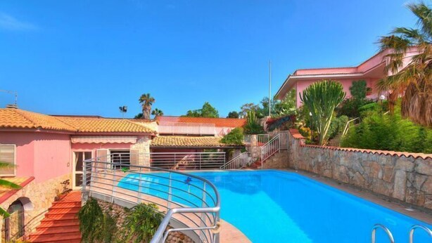 A swimming pool with a stone wall and metal railing, surrounded by a pink building with a tiled roof and a staircase leading to a door. - Holiday Home in Sicily near Fontane Bianche (Terrauzza)