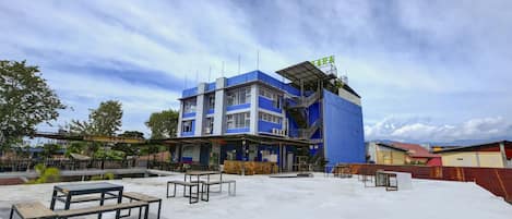 A multi-story building with a blue facade, outdoor seating area with tables and benches, and a clear sky.