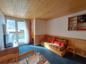 Ein gemütliches Zimmer mit Holzbalkendecke, einem Sofa mit bunten Kissen, einem Fernseher auf einem Holzgestell und einem Esstisch mit Stühlen.
