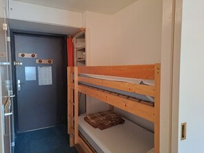 Ein Stockbett mit Holzrahmen und einer Matratze, eine Tür mit Griff und einem Hinweis sowie ein an der Wand befestigtes Regal mit einem weißen Gegenstand.