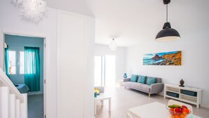 A bright living room with a sofa, a dining table with fruit, and a painting of a coastal scene.