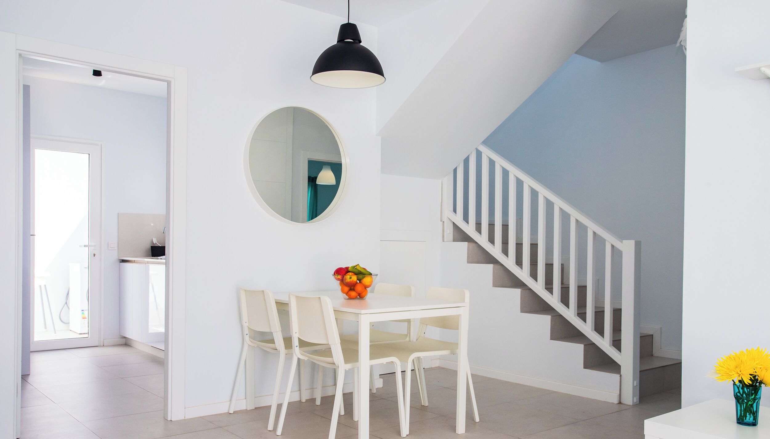 A dining area with a white table and chairs, a round mirror on the wall, and a staircase with white railings.