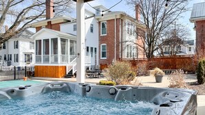 A hot tub in a backyard with a wooden deck, a covered patio, and a brick house in the background. - Beekeeper's Cottage - Hot Tub, Heated Pool, Dog-friendly, Downtown (Washington)
