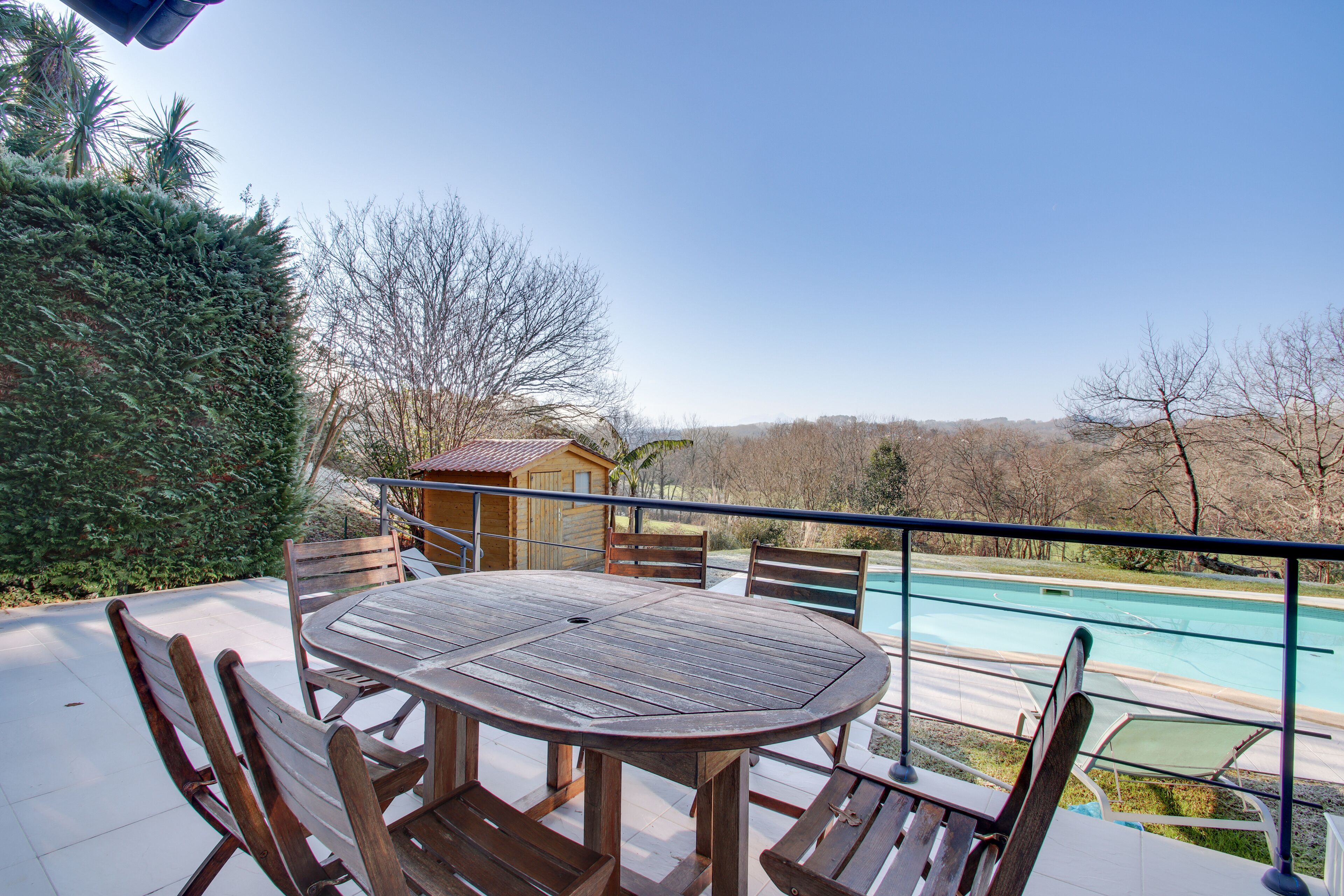 Une table et des chaises en bois sur un balcon donnant sur une piscine et le paysage environnant.