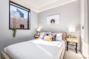 Un dormitorio ordenado con una cama grande, dos mesitas de noche con lámparas, una ventana con vistas a edificios y un cuadro en la pared.