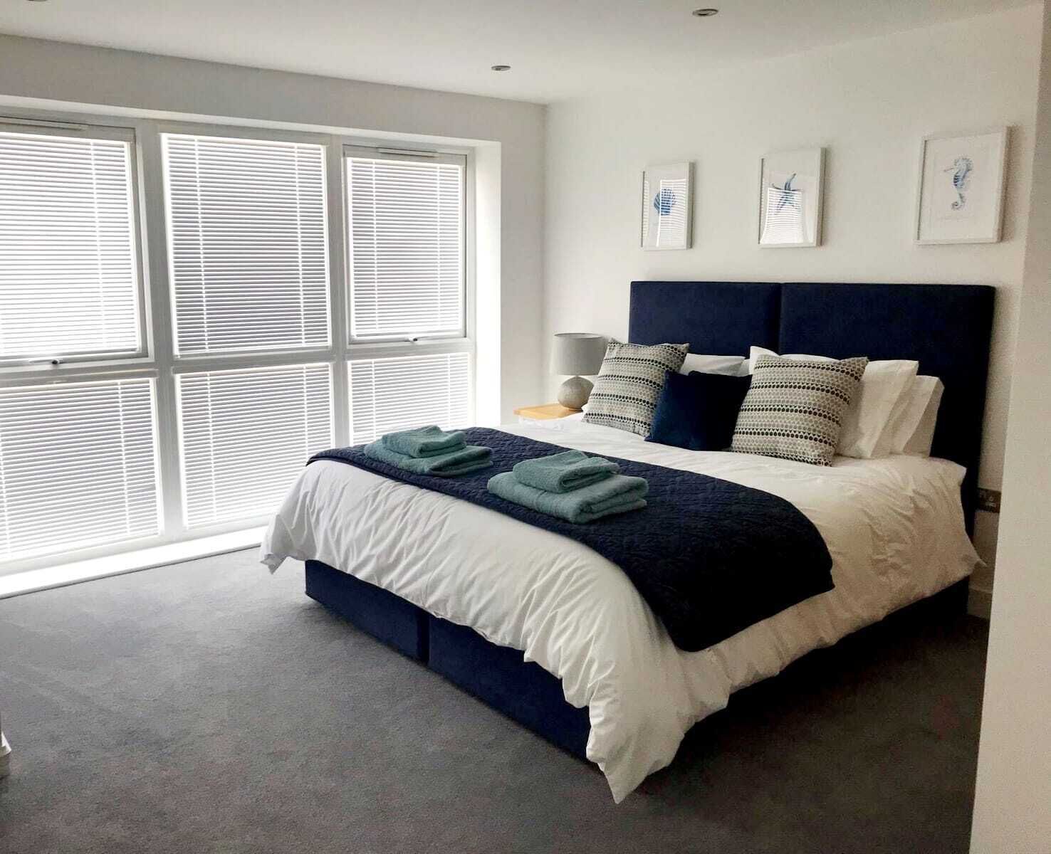 A modern bedroom with a large bed, white bedding, and a dark blue headboard. There are three framed artworks on the wall and a window with blinds.