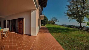 Ein überdachter Patio-Bereich mit Esstisch und Stühlen, einem gefliesten Boden und Blick auf eine grüne Landschaft mit Bäumen und einem Zaun.