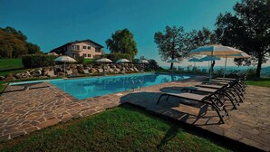 Ein Swimmingpool mit Liegestühlen und Sonnenschirmen, umgeben von einer Steinterrasse und einem großen Haus im Hintergrund.