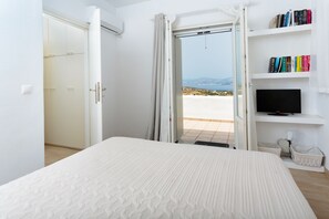 A bedroom with a bed, a television, a bookshelf, and a door leading to a balcony with a view.