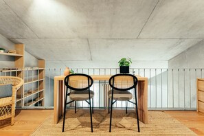 Ett rum i loftstil med ett betongtak, ett matbord i trä med två stolar, en fåtölj i rotting, en bokhylla och en krukväxt.
