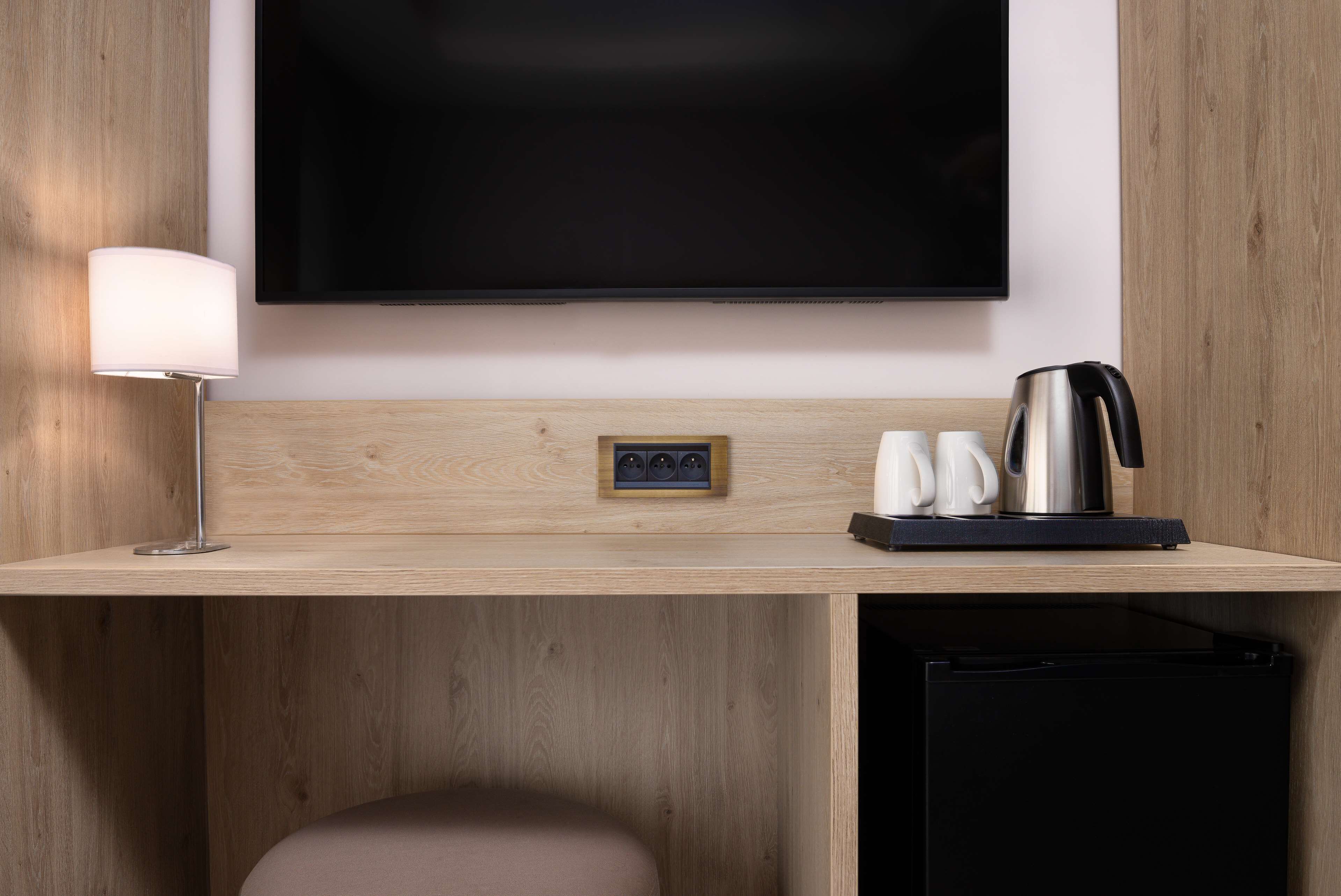 A hotel room with a wooden desk, a flat-screen TV, a lamp, a kettle, and a small refrigerator.