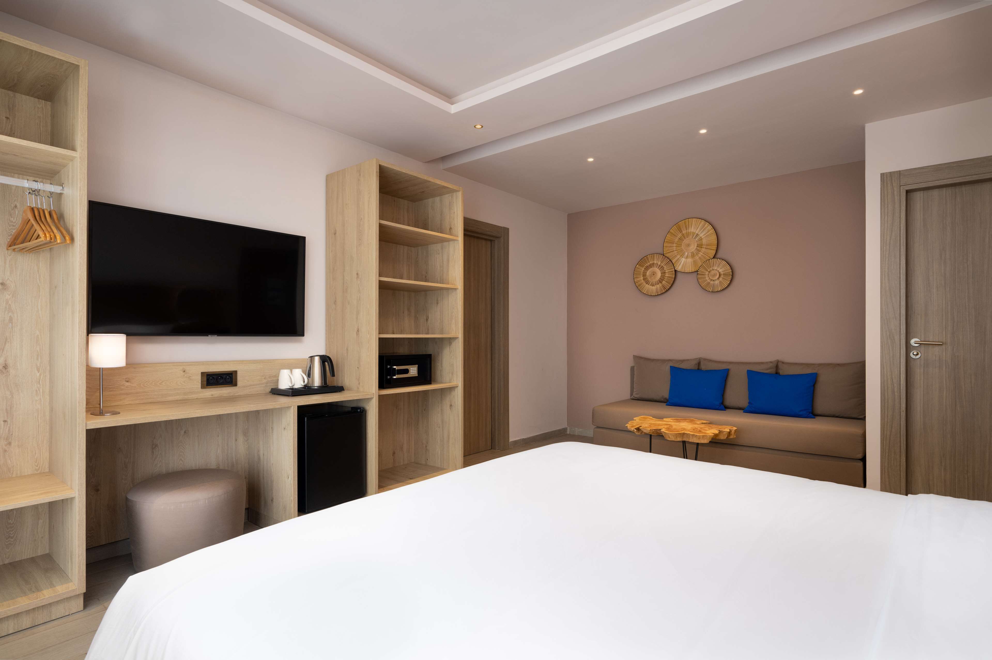 A modern hotel room with a large bed, a TV mounted on a wooden wall, a seating area with a sofa, and built-in storage units.