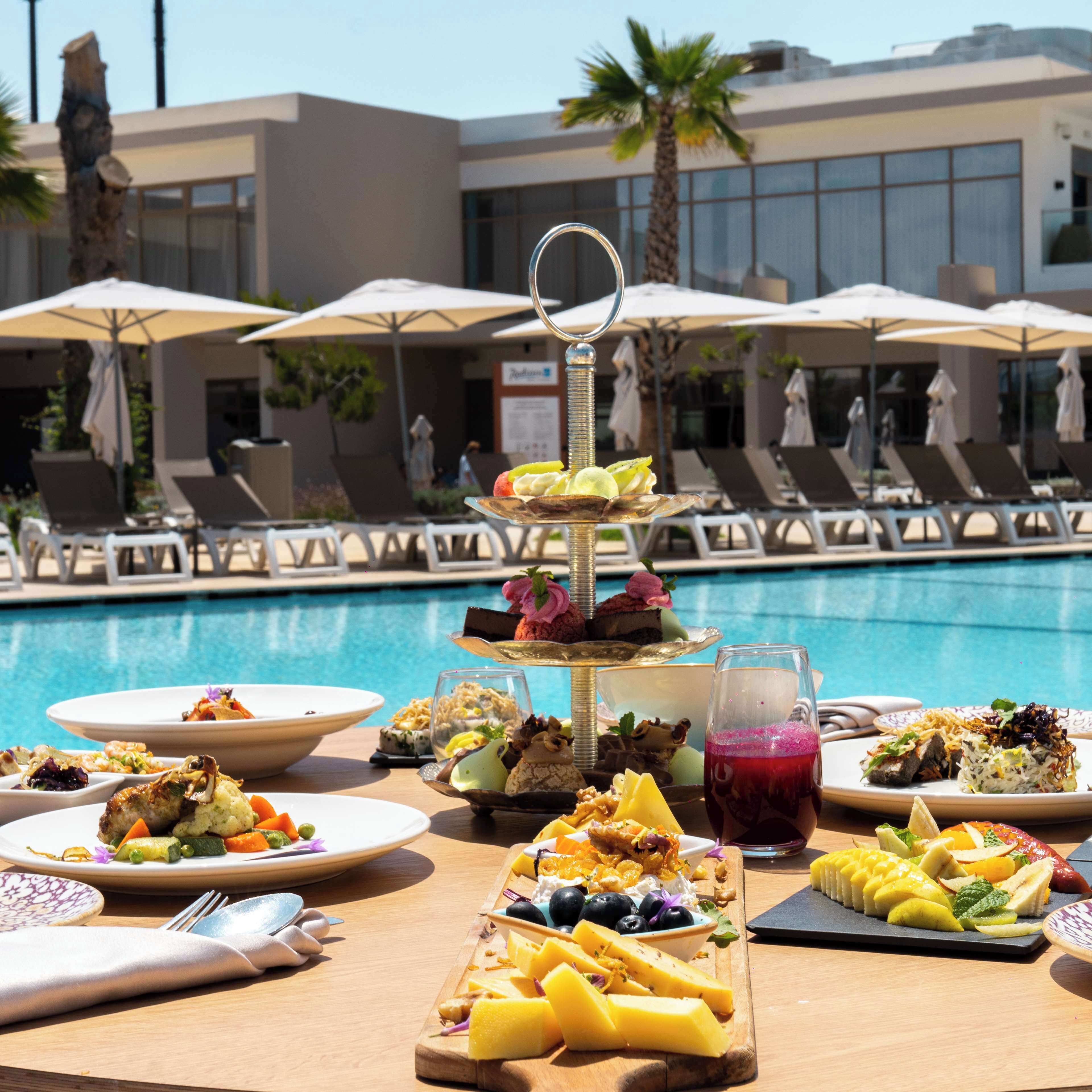 A poolside buffet with various dishes and a drink, surrounded by lounge chairs and umbrellas.