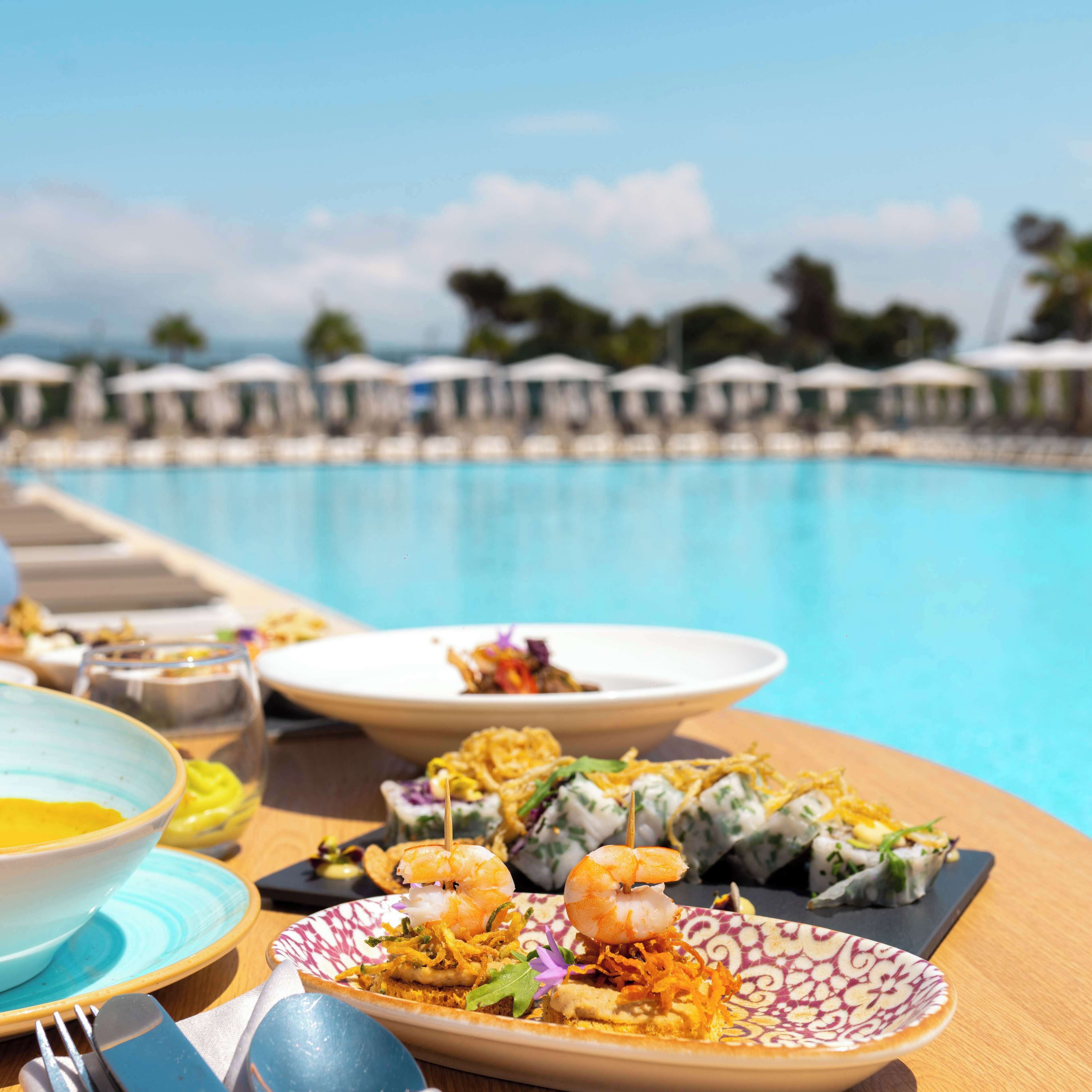 A dining table with plates of food, a pool, and umbrellas in the background.