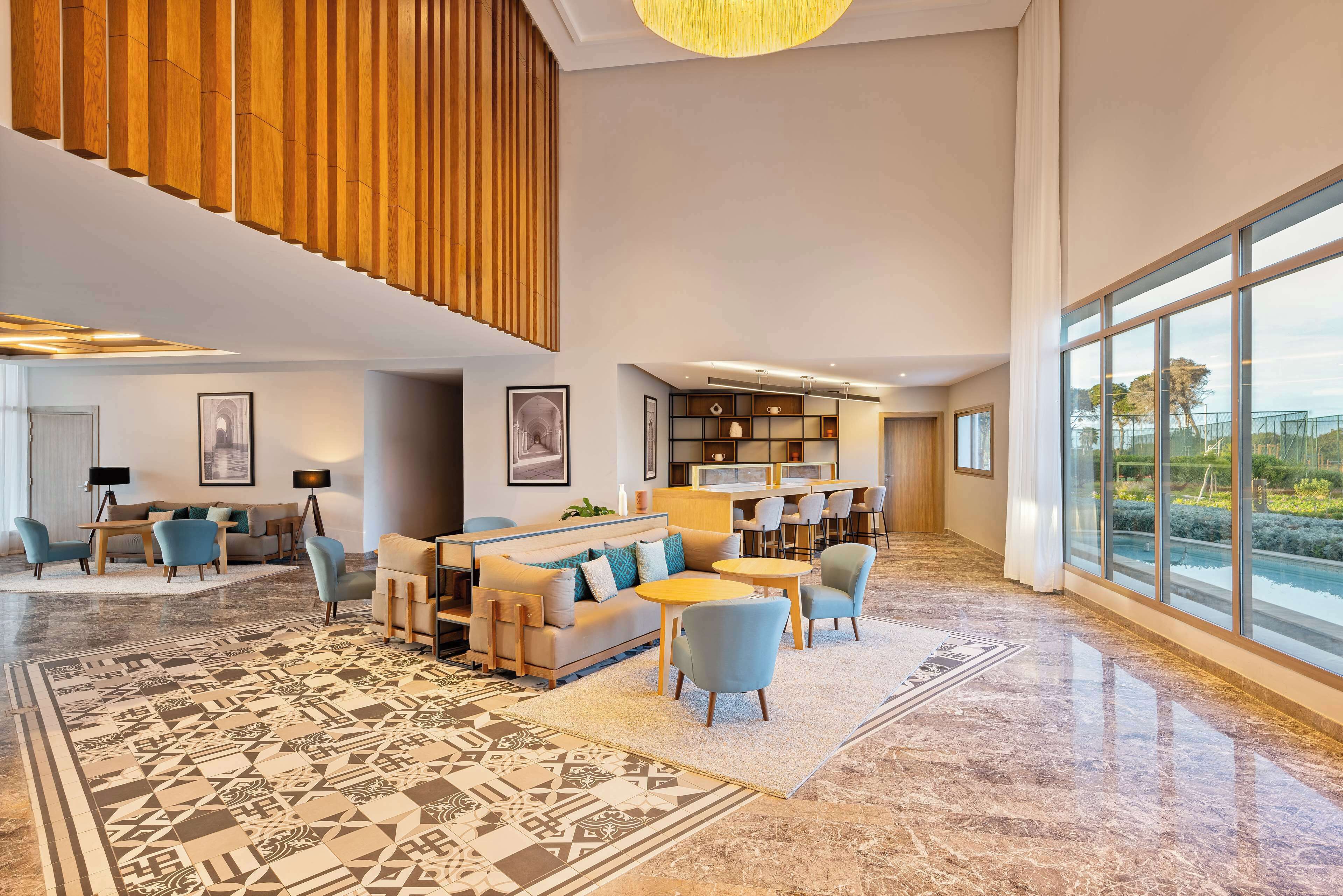 A spacious lobby with a high ceiling, a large window offering a view of the outdoors, a seating area with sofas and chairs, and a patterned floor.