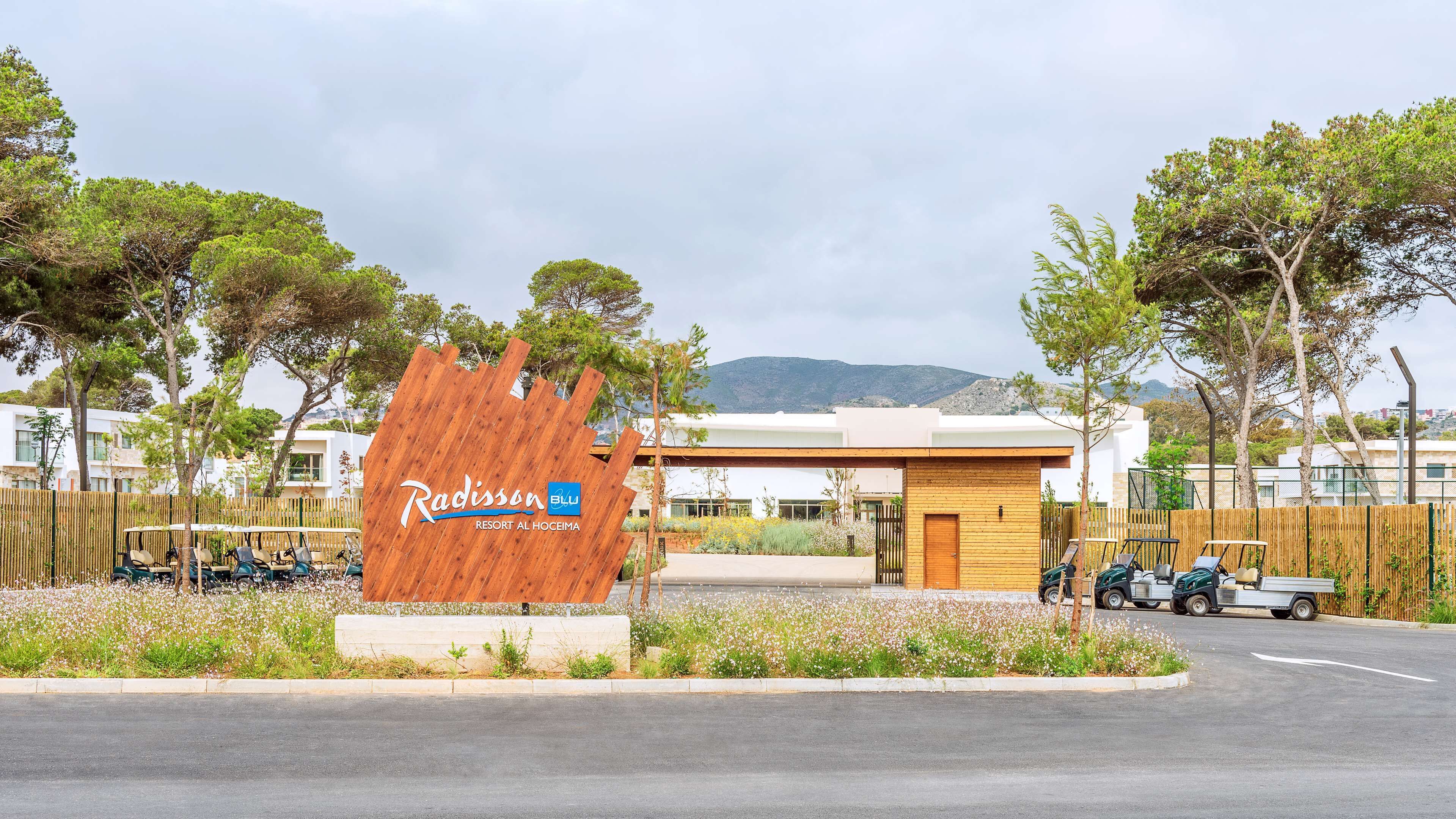 A resort entrance with a wooden sign, a parking area, and a view of buildings and mountains in the background.