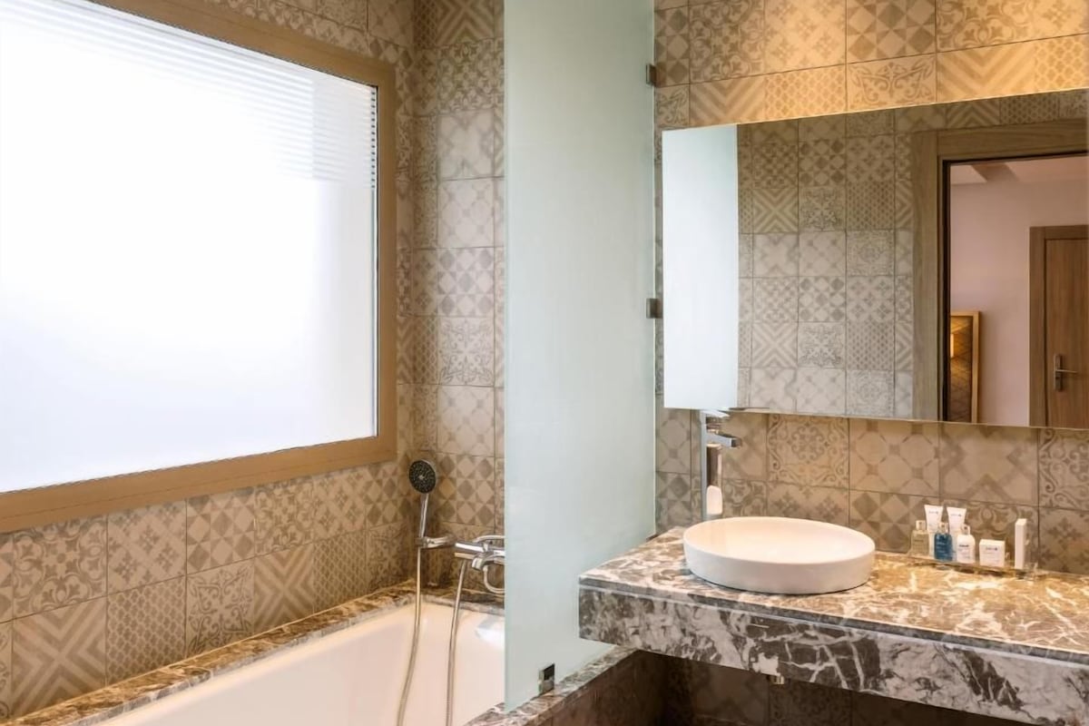 A bathroom with patterned tiles, a bathtub, a shower, a sink with toiletries, and a mirror.