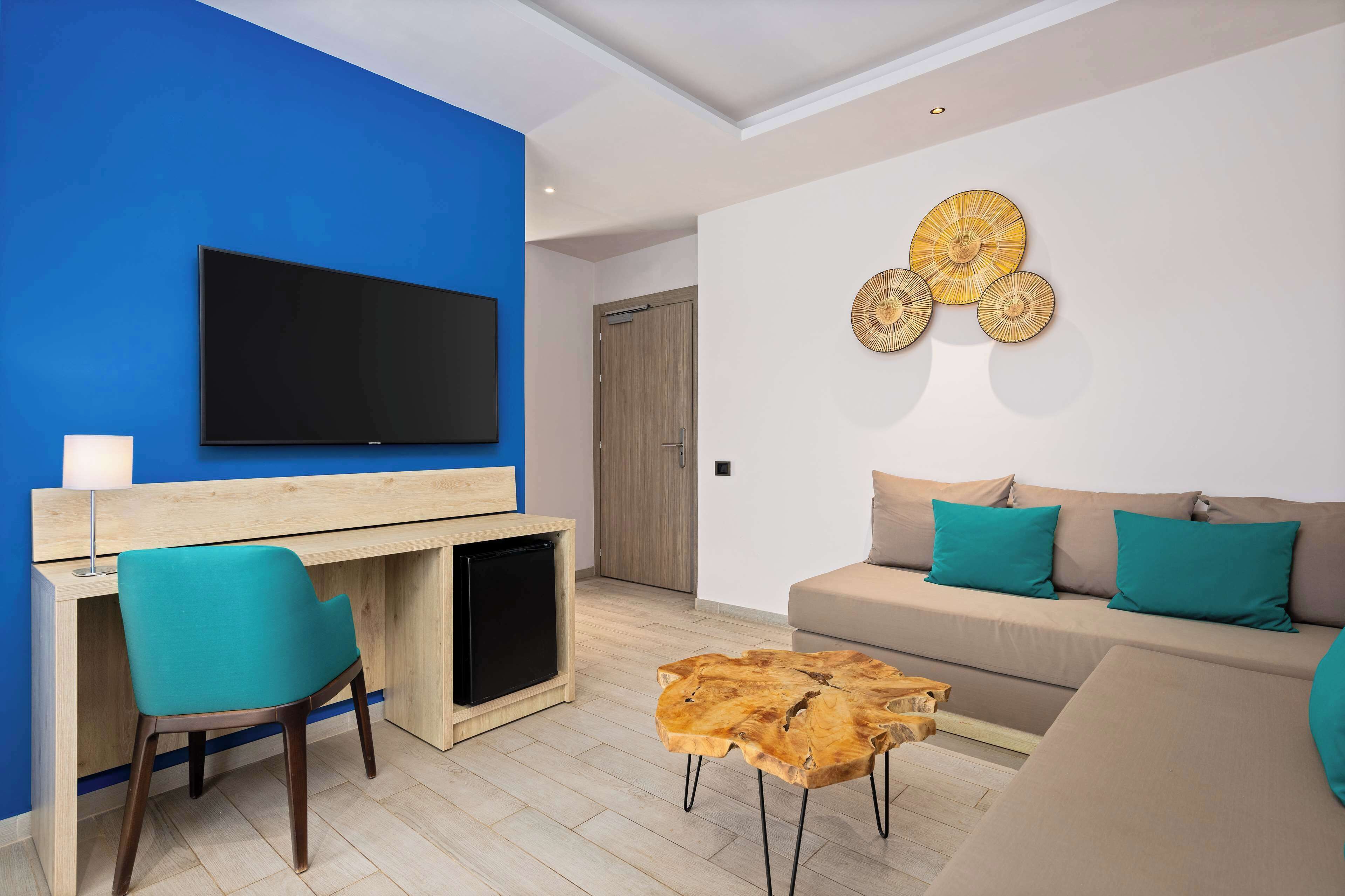 Ein modernes Wohnzimmer mit blauer Akzentwand, einem Flachbildfernseher, einem Holztisch, einem türkisfarbenen Sessel, einem beige gefärbten Sofa mit türkisfarbenen Kissen und einem Couchtisch aus Holz.