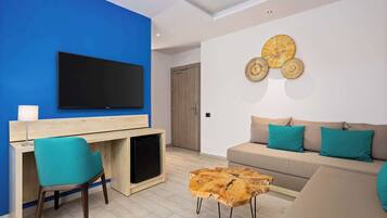 Ein modernes Wohnzimmer mit blauer Akzentwand, einem Flachbildfernseher, einem Holztisch, einem türkisfarbenen Sessel, einem beige gefärbten Sofa mit türkisfarbenen Kissen und einem Couchtisch aus Holz.