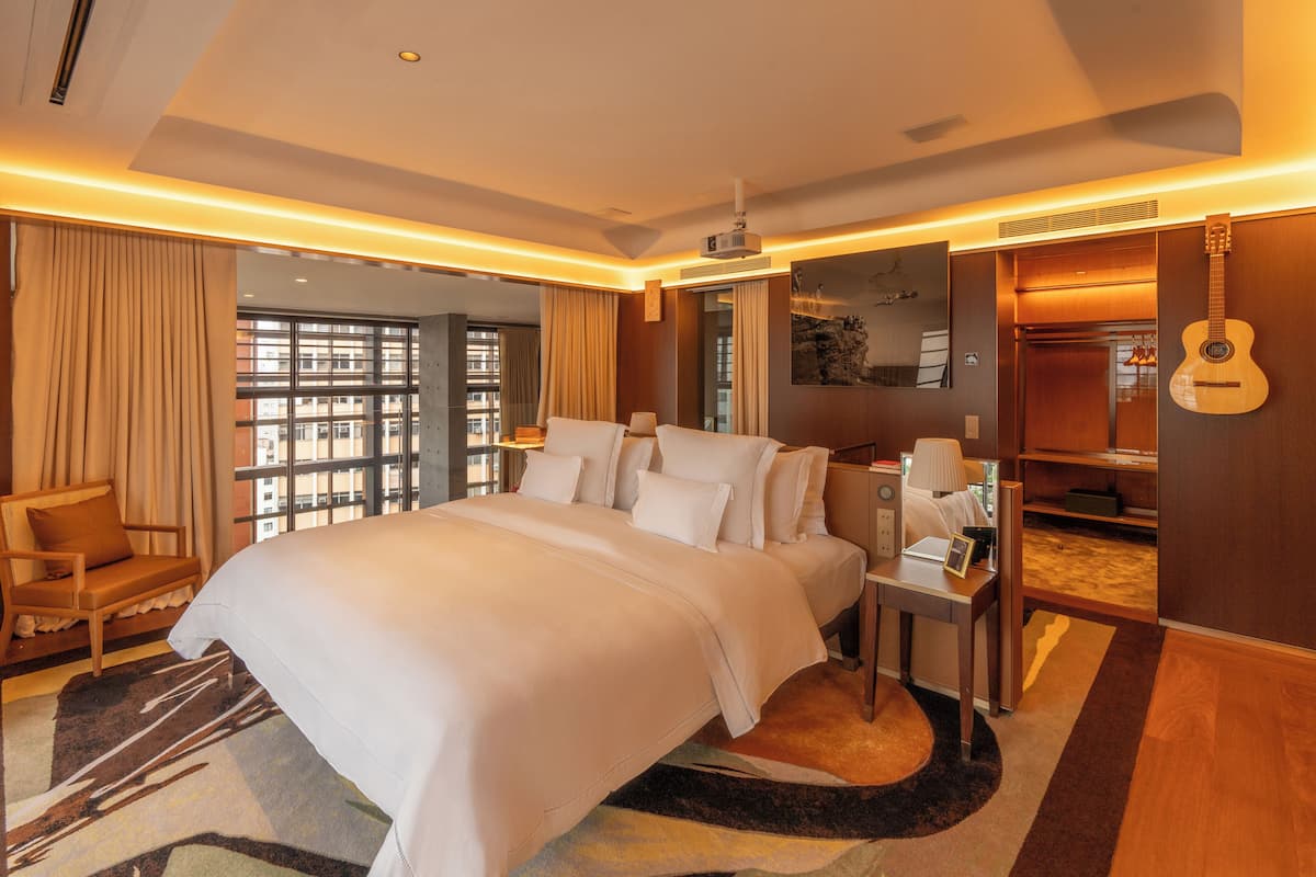 Ein geräumiges Hotelzimmer mit einem großen Bett, einem Schreibtisch, einem Stuhl, einer Gitarre und Blick auf die Stadt durch große Fenster.