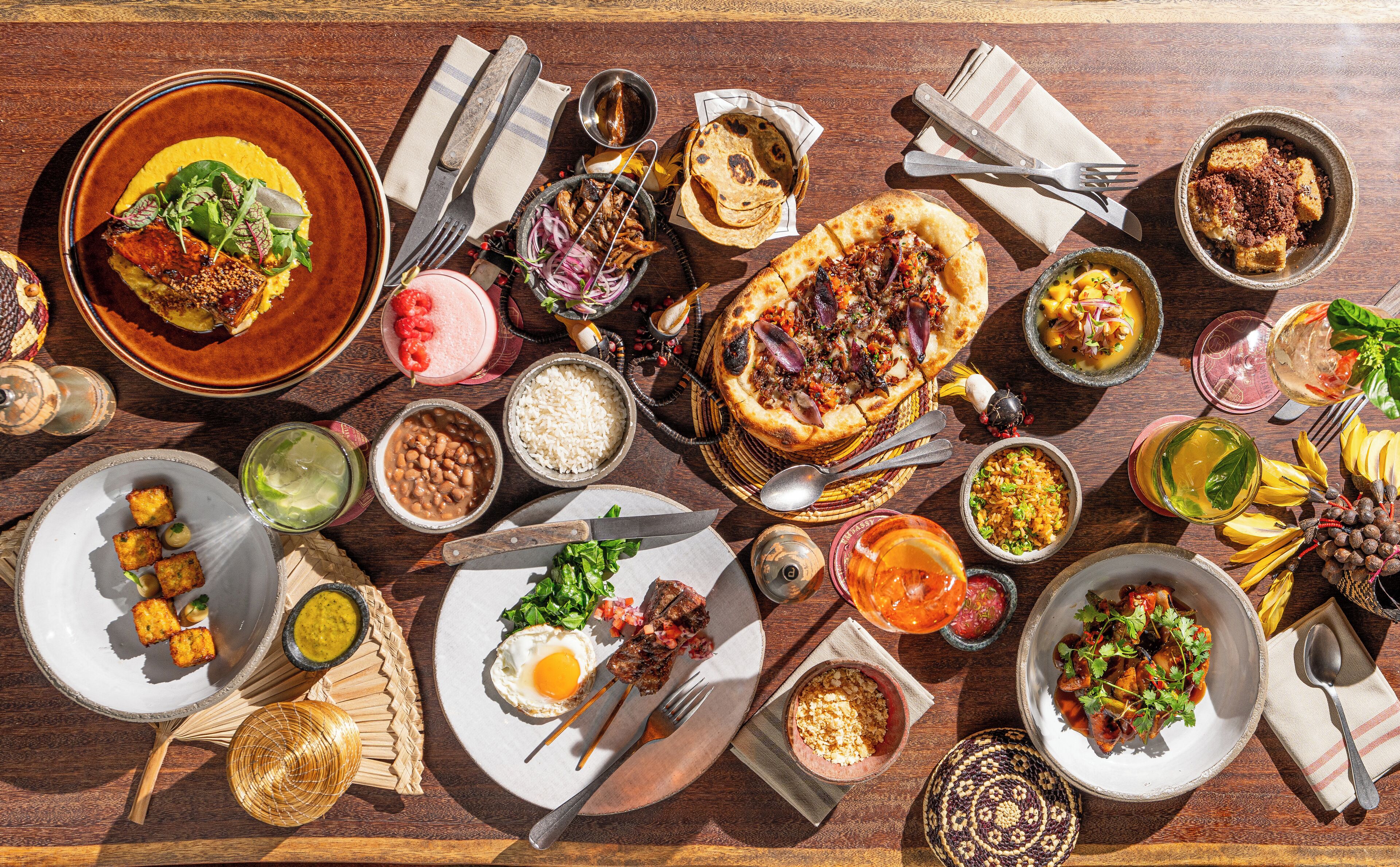 Diversos pratos, incluindo pizza, arroz, feijão e um ovo frito, sobre uma mesa de madeira.