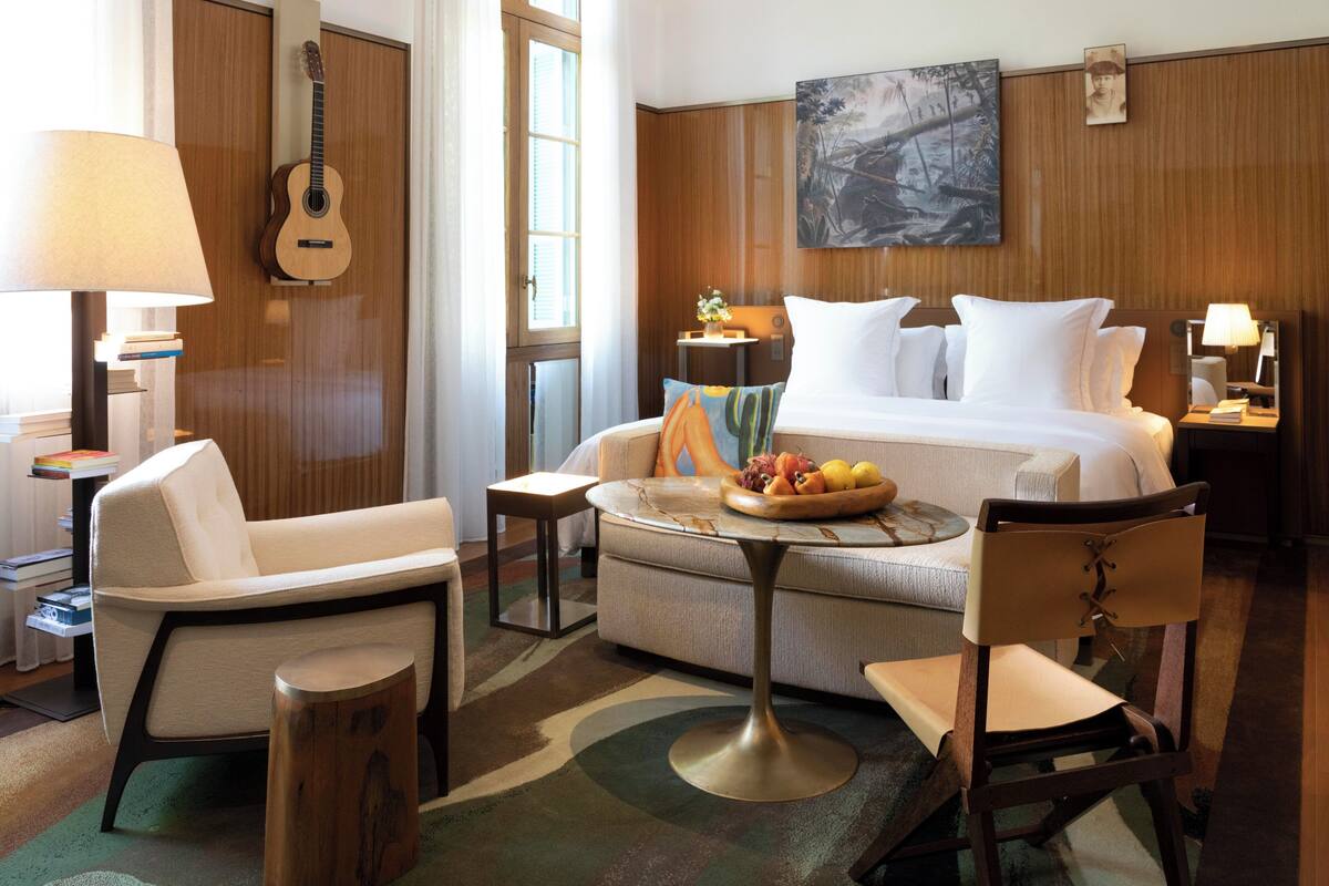 Ein Hotelzimmer mit einem Bett, einer Couch, einem Sessel, einem kleinen Tisch mit Obst, einer Lampe, einer Gitarre und einem Bild an der Wand.
