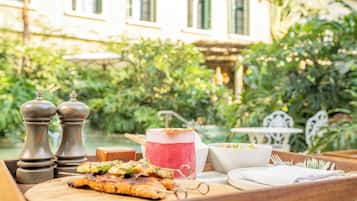 A wooden tray with grilled food, a cocktail, and salt and pepper shakers on a patio with a building and greenery in the background.
