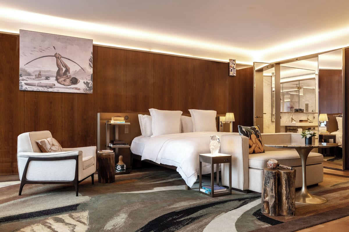 Ein modernes Hotelzimmer mit einem großen Bett, einer Sitzecke mit Stuhl und kleinem Tisch, einem großen Spiegel und einem Bild an der Wand.