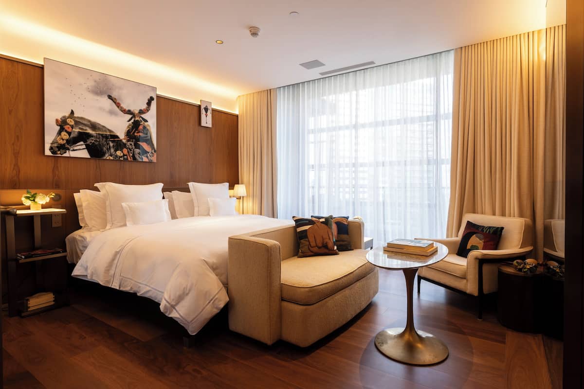 Ein modernes Hotelzimmer mit einem großen Bett, einer Couch, einem kleinen Tisch und einem Sessel. An der Wand hängt ein Gemälde und ein Fenster mit Vorhängen.