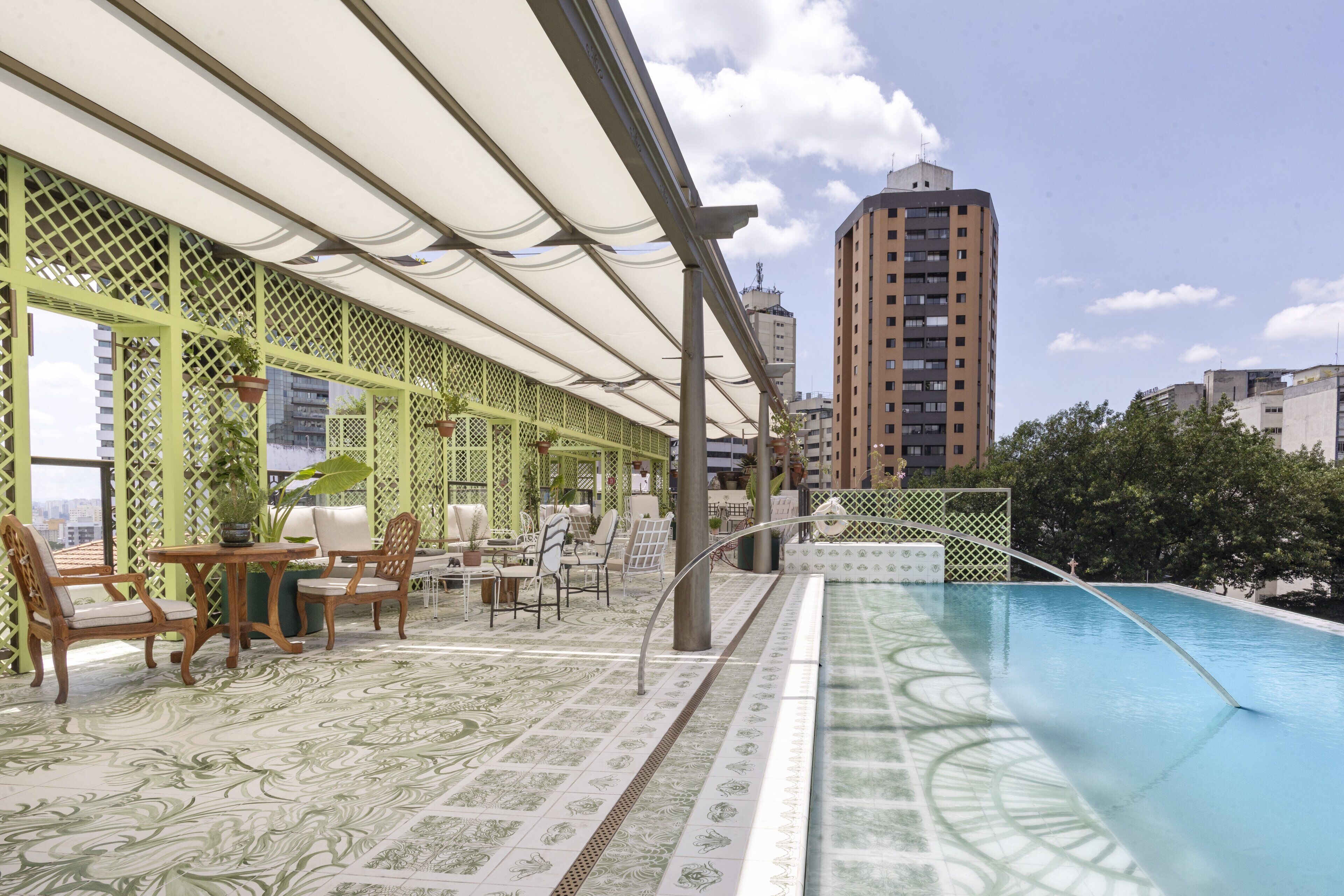 A rooftop pool area with a green lattice fence, outdoor seating, and a view of tall buildings.