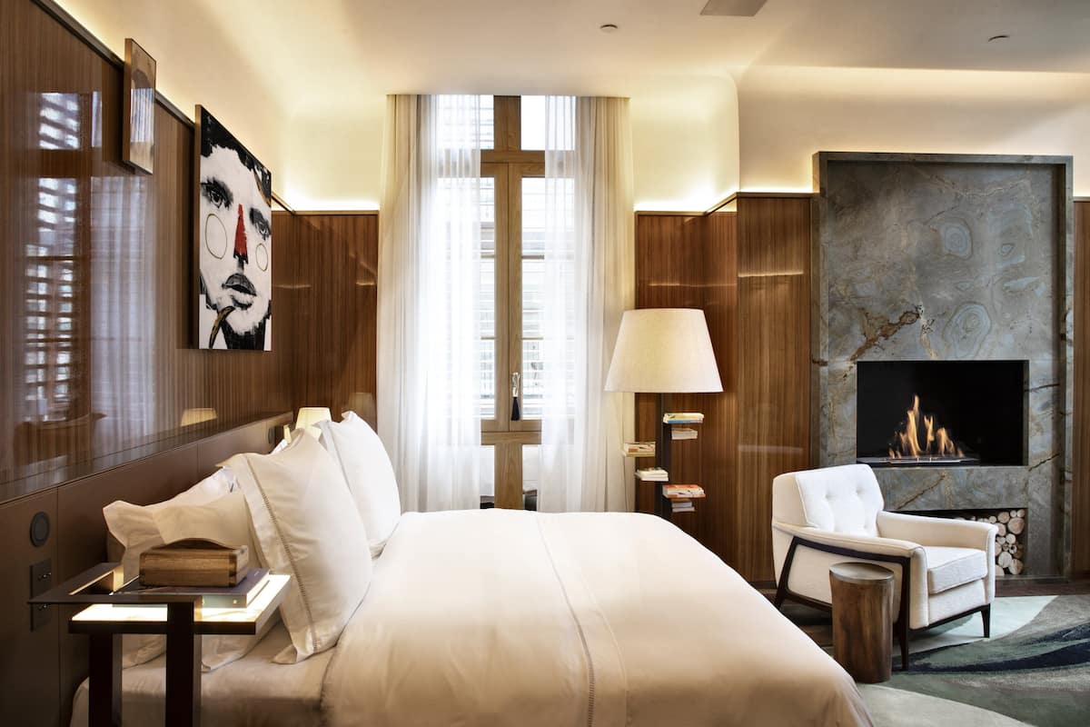 Ein modernes Hotelzimmer mit einem großen Bett, einer Sitzecke mit einem Sessel, einem Kamin und einem großen Fenster mit durchsichtigen Vorhängen.