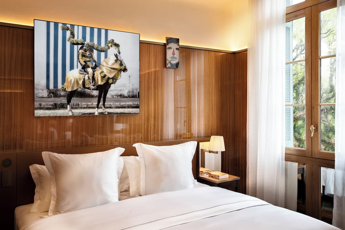Ein Hotelzimmer mit einem großen Bett, einem Nachttisch mit Lampe, einem Gemälde von Reiter und Pferd sowie einem Fenster mit Vorhängen.