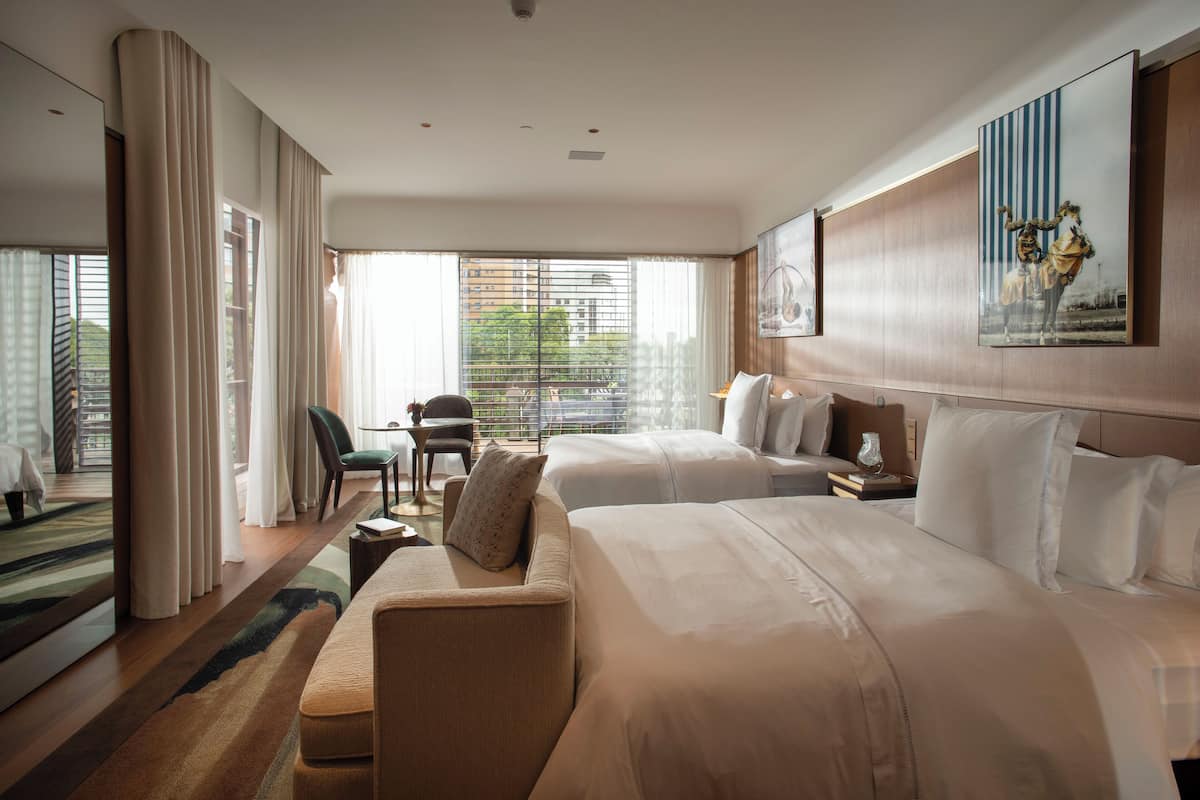 Ein Hotelzimmer mit zwei Betten, einem Sofa, einem Schreibtisch und Blick auf die Stadt durch große Fenster.
