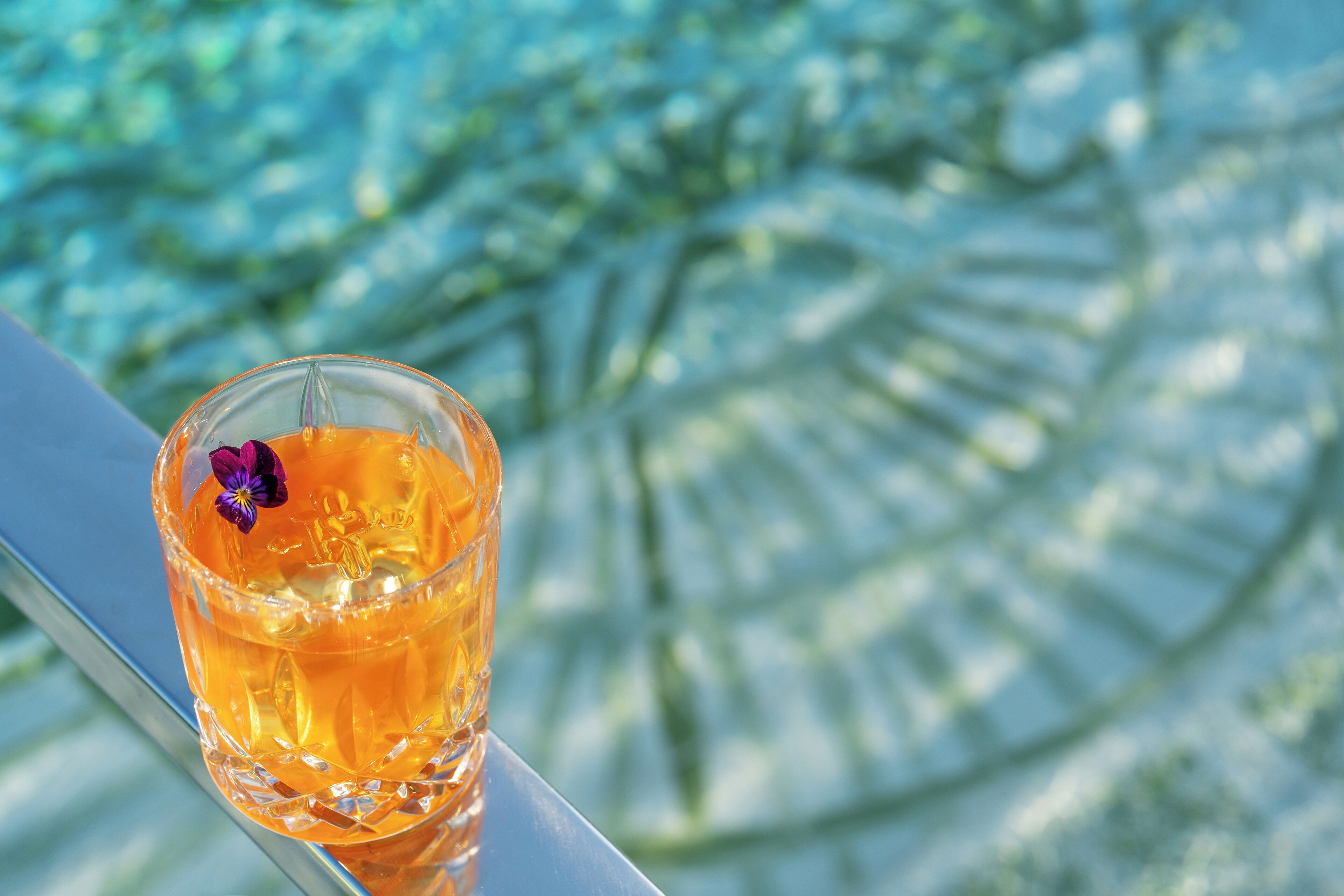 Een glas amberkleurige vloeistof met een paarse bloem op een reflecterend oppervlak.
