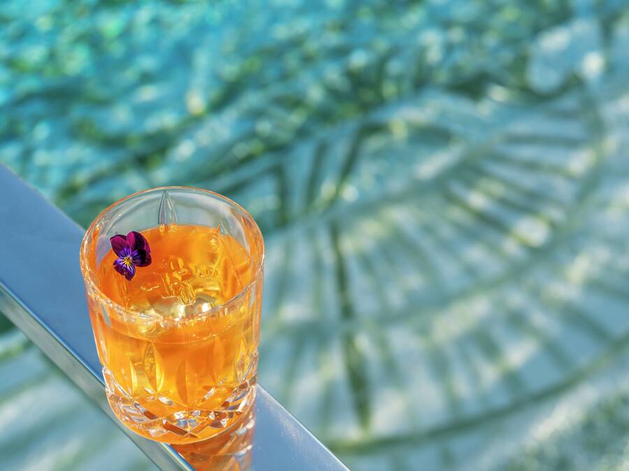Een glas amberkleurige vloeistof met een paarse bloem op een reflecterend oppervlak.