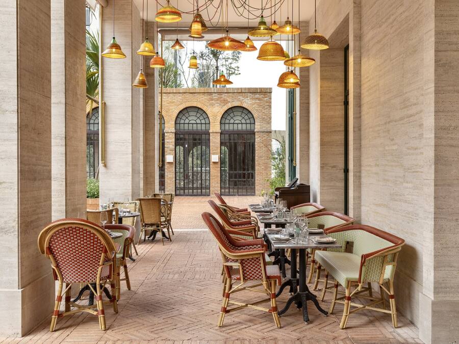 Un comedor interior con sillas y mesas de mimbre, pared de ladrillo y lámparas colgantes.