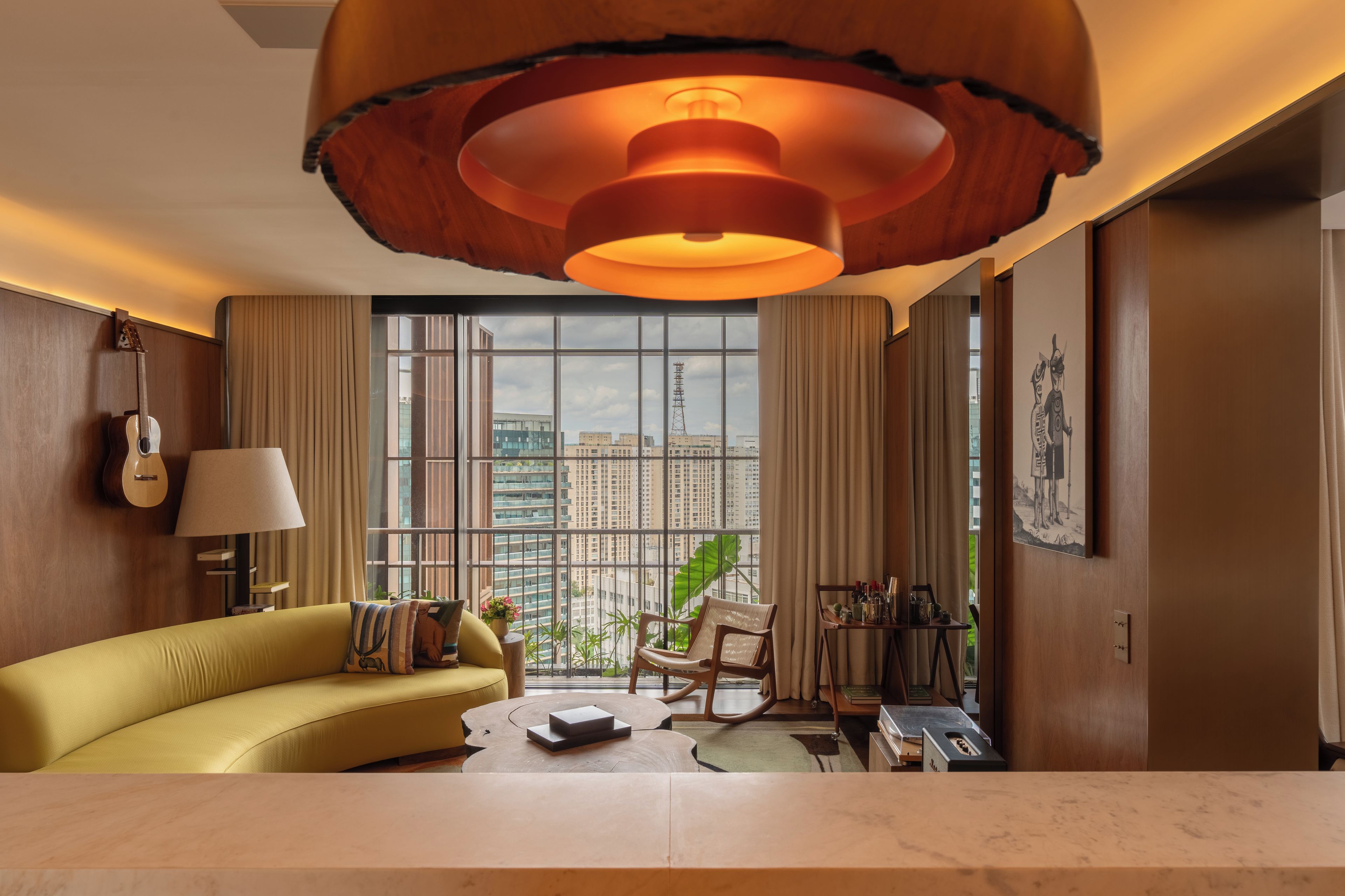 Uma sala de estar moderna com um sofá amarelo, uma janela grande com vista para prédios, uma luminária pendente e uma guitarra na parede.