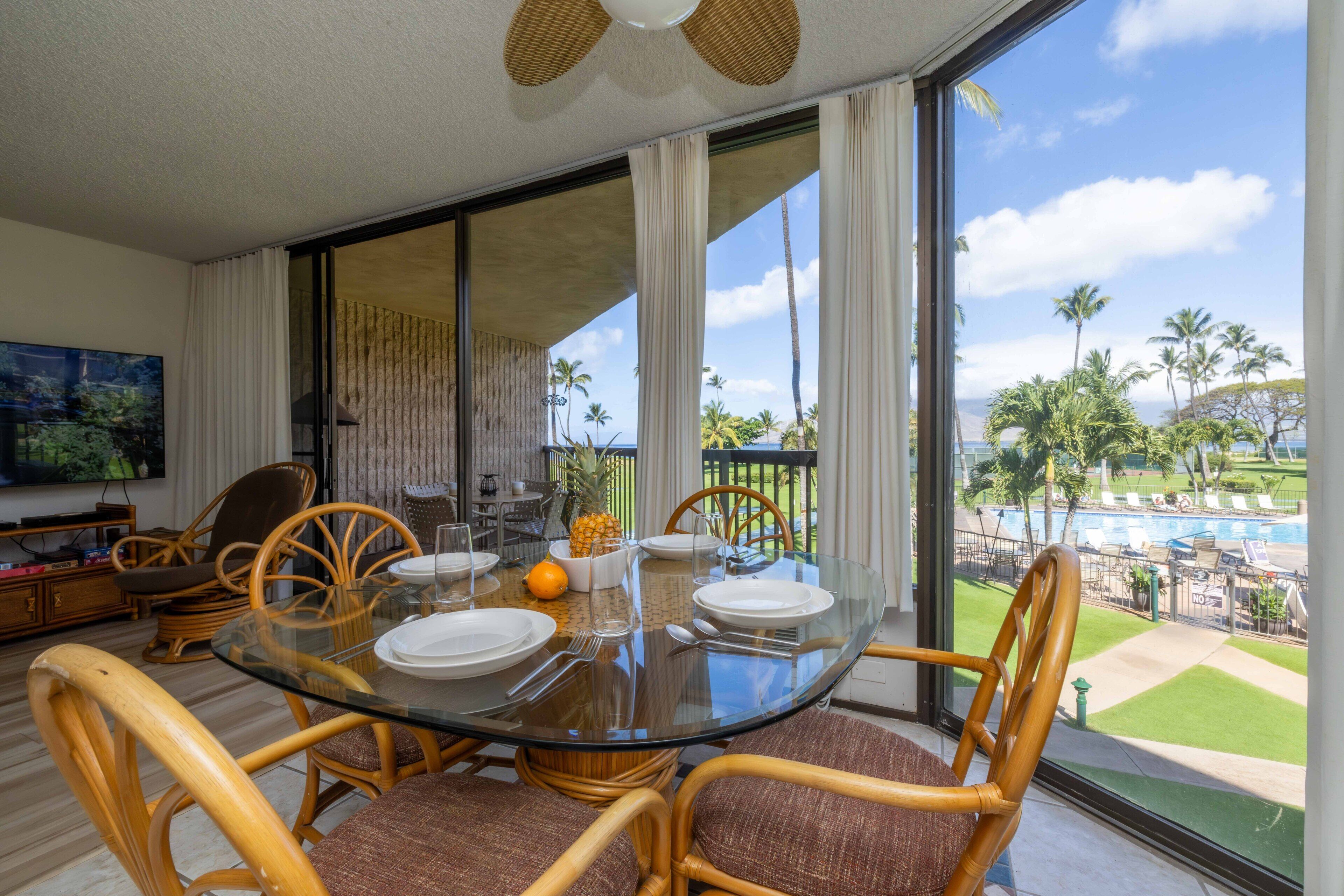 A dining area with a glass table, wicker chairs, and a view of a pool and palm trees.