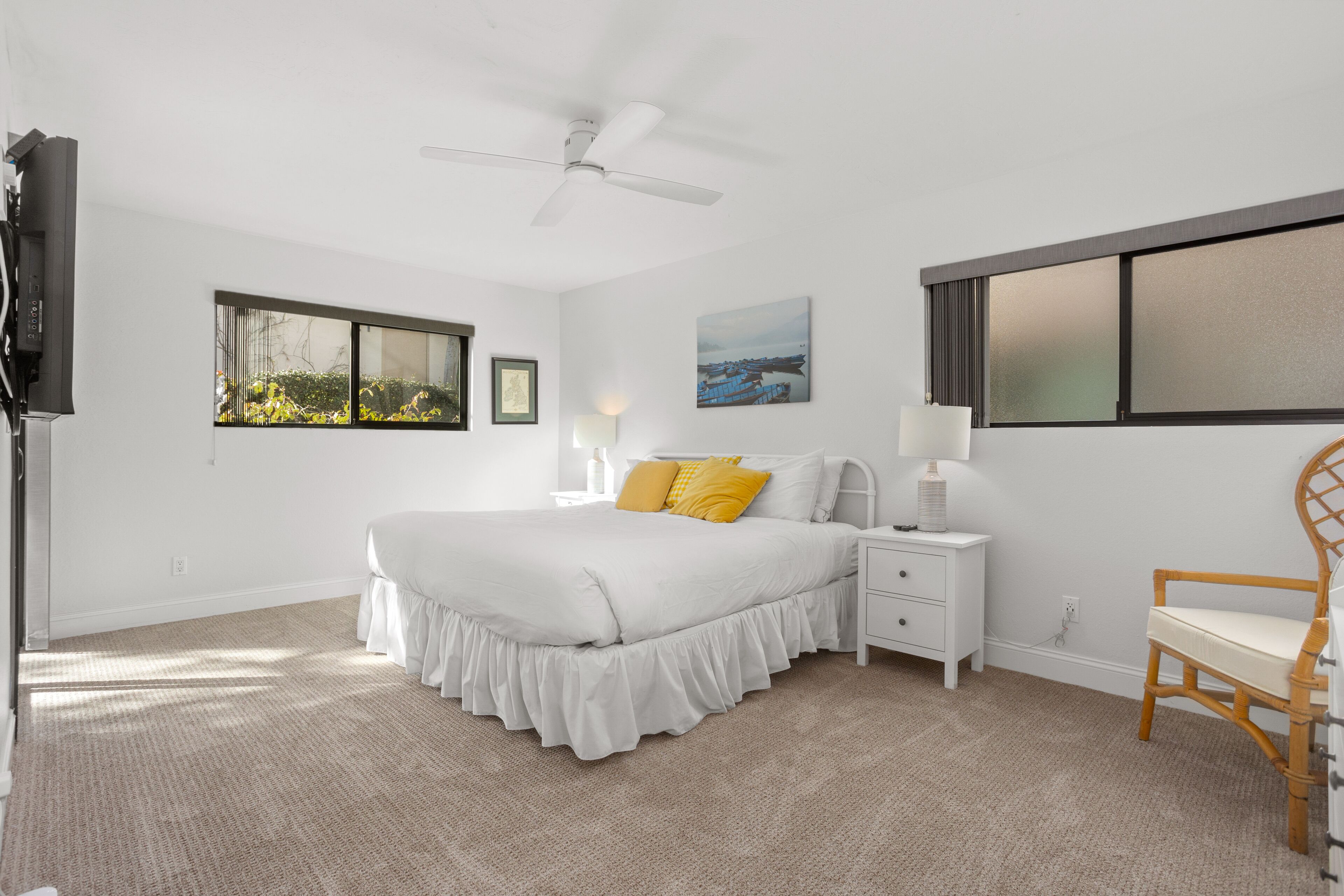 Une chambre à coucher comprenant un lit, une table de chevet, une chaise, un ventilateur de plafond, une fenêtre et un tableau accroché au mur.