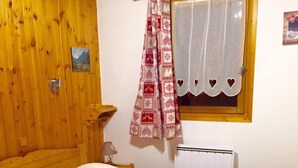 Une pièce chaleureuse avec un mur en bois, un lit recouvert d’une literie blanche, une fenêtre ornée d’un rideau à motifs en forme de cœur et un radiateur.