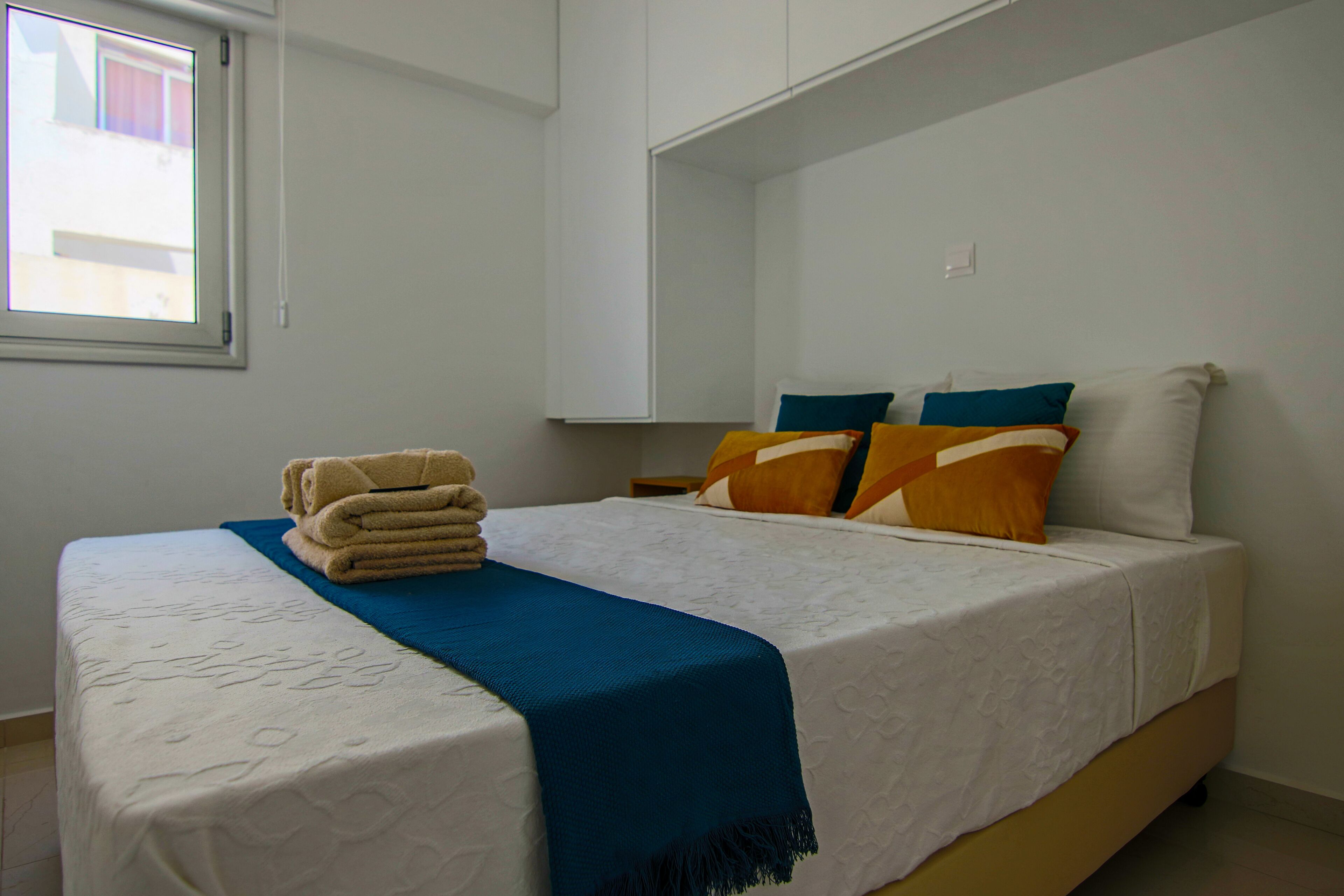 A neatly made bed with a blue throw blanket and a stack of towels, white bedding, and two pillows with geometric patterns.