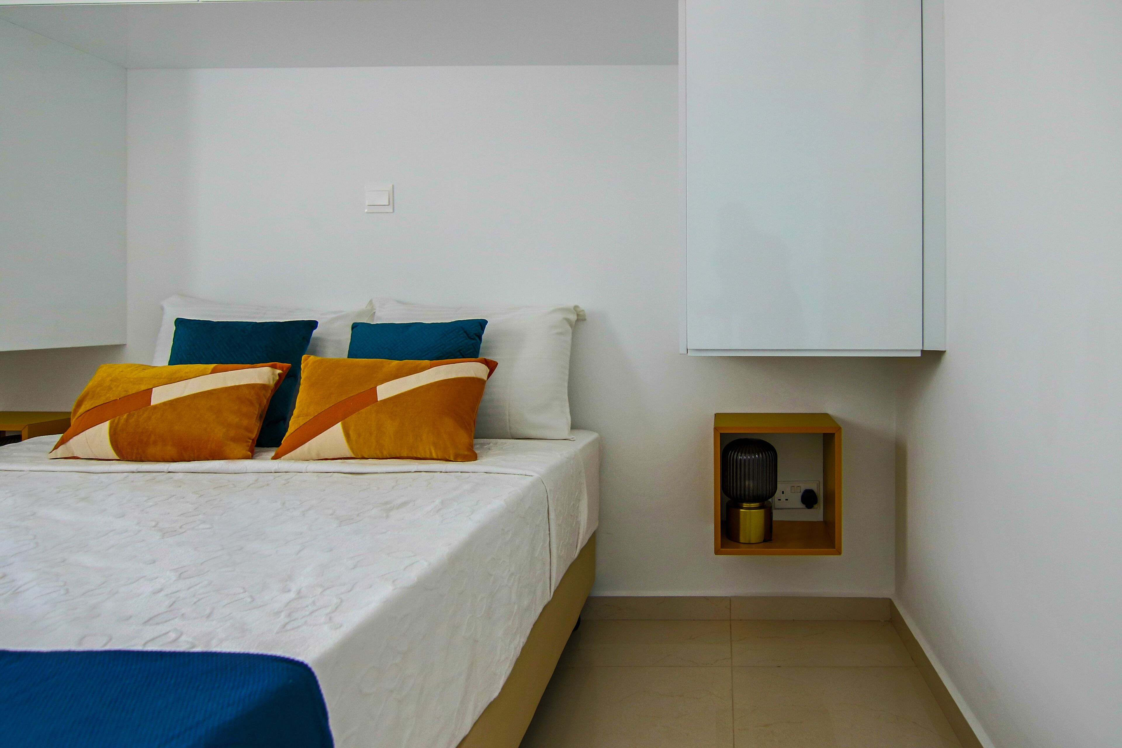 A neatly made bed with a white mattress and multiple pillows, a small wooden nightstand with a lamp, and a white wall with a light switch.