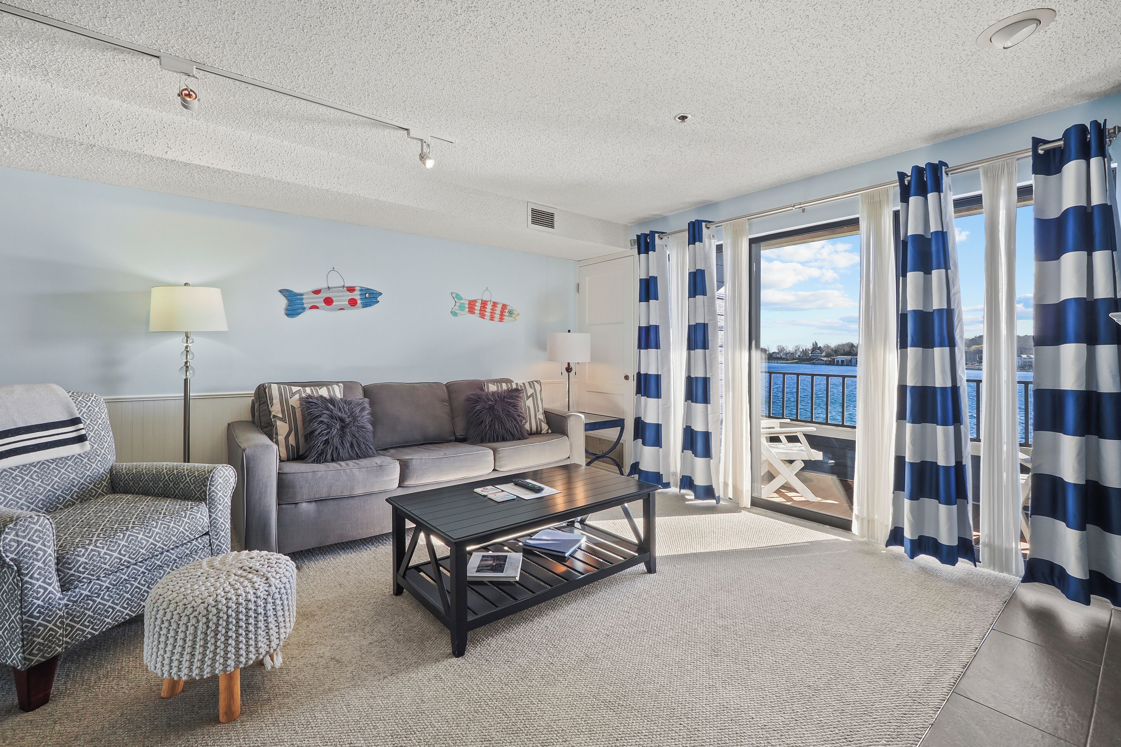 A living room with a grey sofa, a patterned armchair, a coffee table, and a small ottoman. There are fish wall decorations and a view of the sea through the curtains.