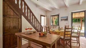 Un coin repas avec une table en bois dressée pour quatre personnes, un panier de fruits et des verres à vin posés sur la table.