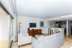 A modern living room with a sofa, a coffee table, and a TV. There is a kitchen area with a refrigerator and a microwave.