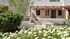 Une cour bordée de bâtiments aux murs blanchis à la chaux, une pergola et un jardin où s’épanouissent des fleurs blanches et jaunes.